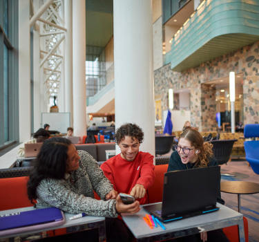 Three sitting students engaged in conversation smiling in front of a laptop.